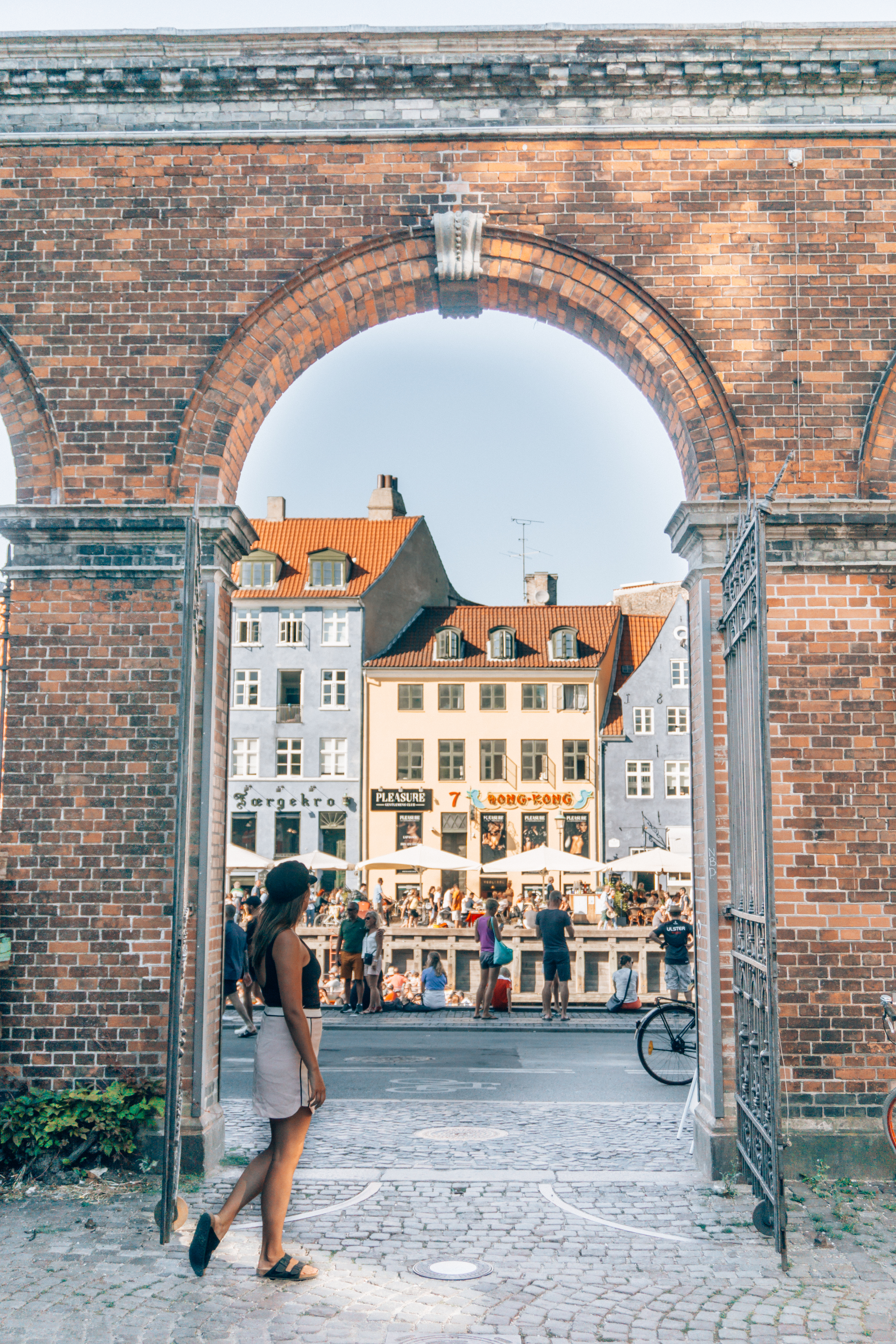 A girl admires the bustling Nyhavn waterfront district in Copenhagen from behind an old brick archway.