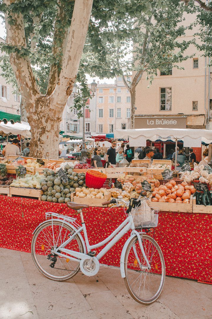 A light blue bike is parked outside the Aix-en-Provence market in the city square, with fruits and vegetables in the background. 
