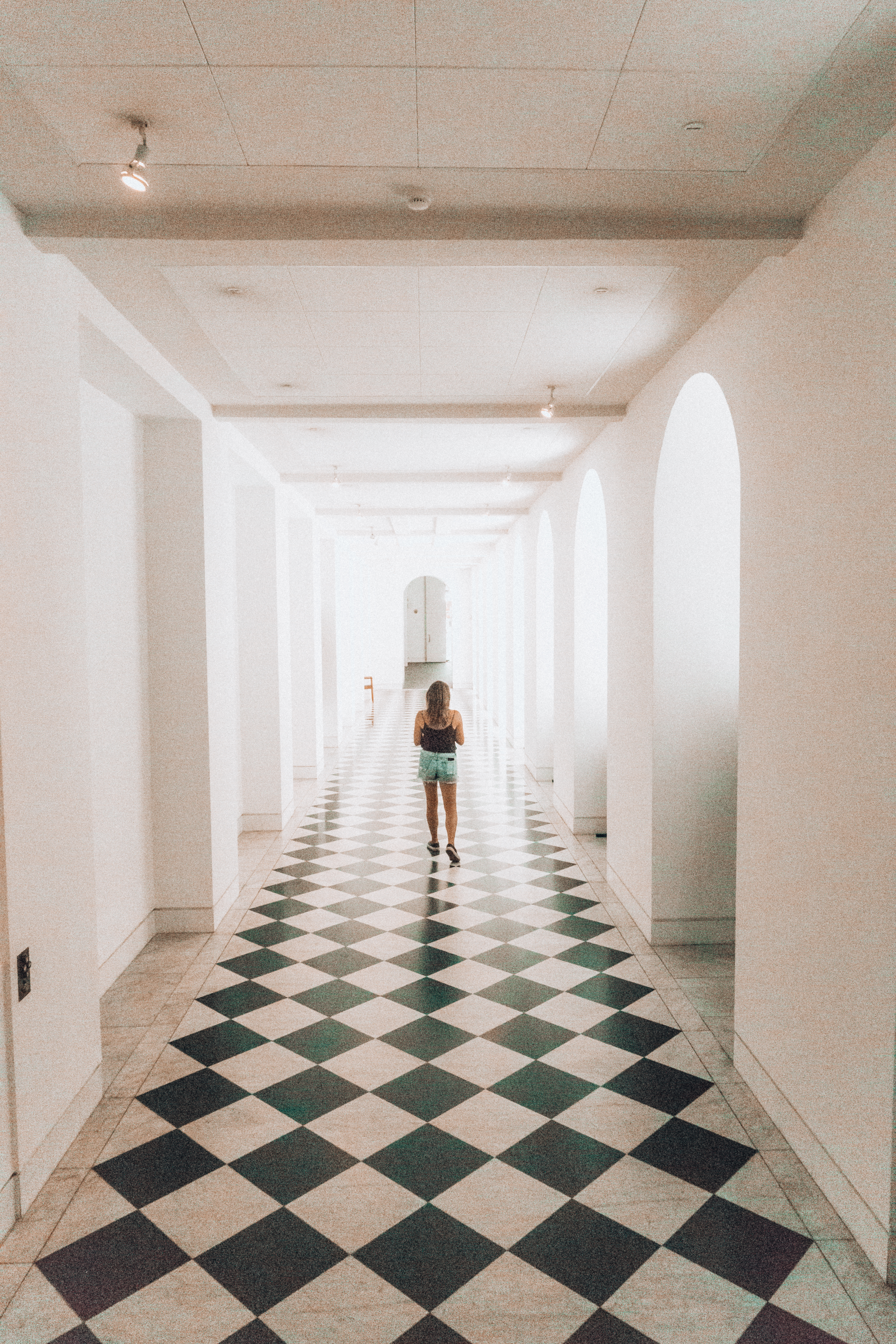 A girl wearing a black top and shorts walks down a long white hallway that has checkered floors and archways