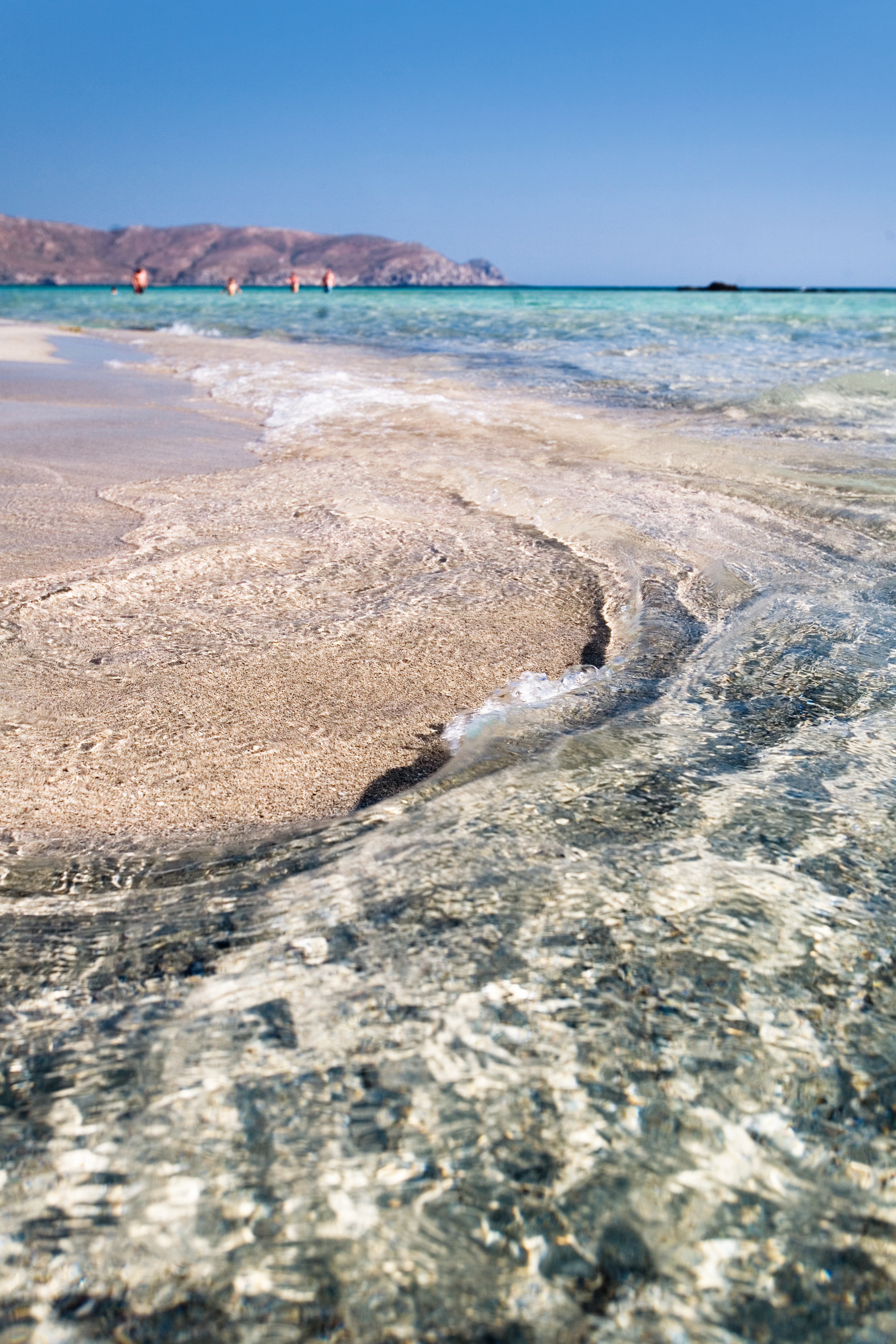 Clear ocean water splashes up onto the iconic pink sand of Elafonisi Beach on Crete Island in Greece with people swimming in the background