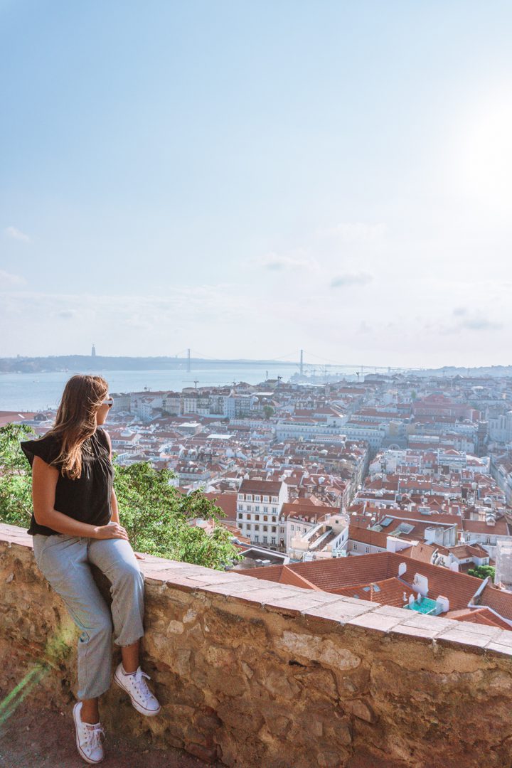 A lady looks out over the sweeping views of red roofs in Lisbon, on a sunny summer day with the Ponte 25 de Abril bridge and River Tagus in the background. 