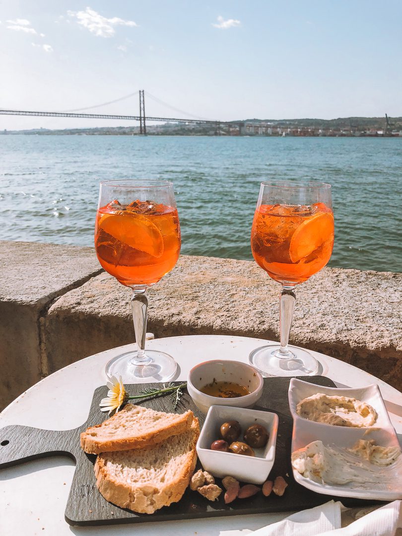 Two aperol spritz' sit on a table with some tapas and the Ponte 25 de Abril bridge and River Tagus in the background. 
