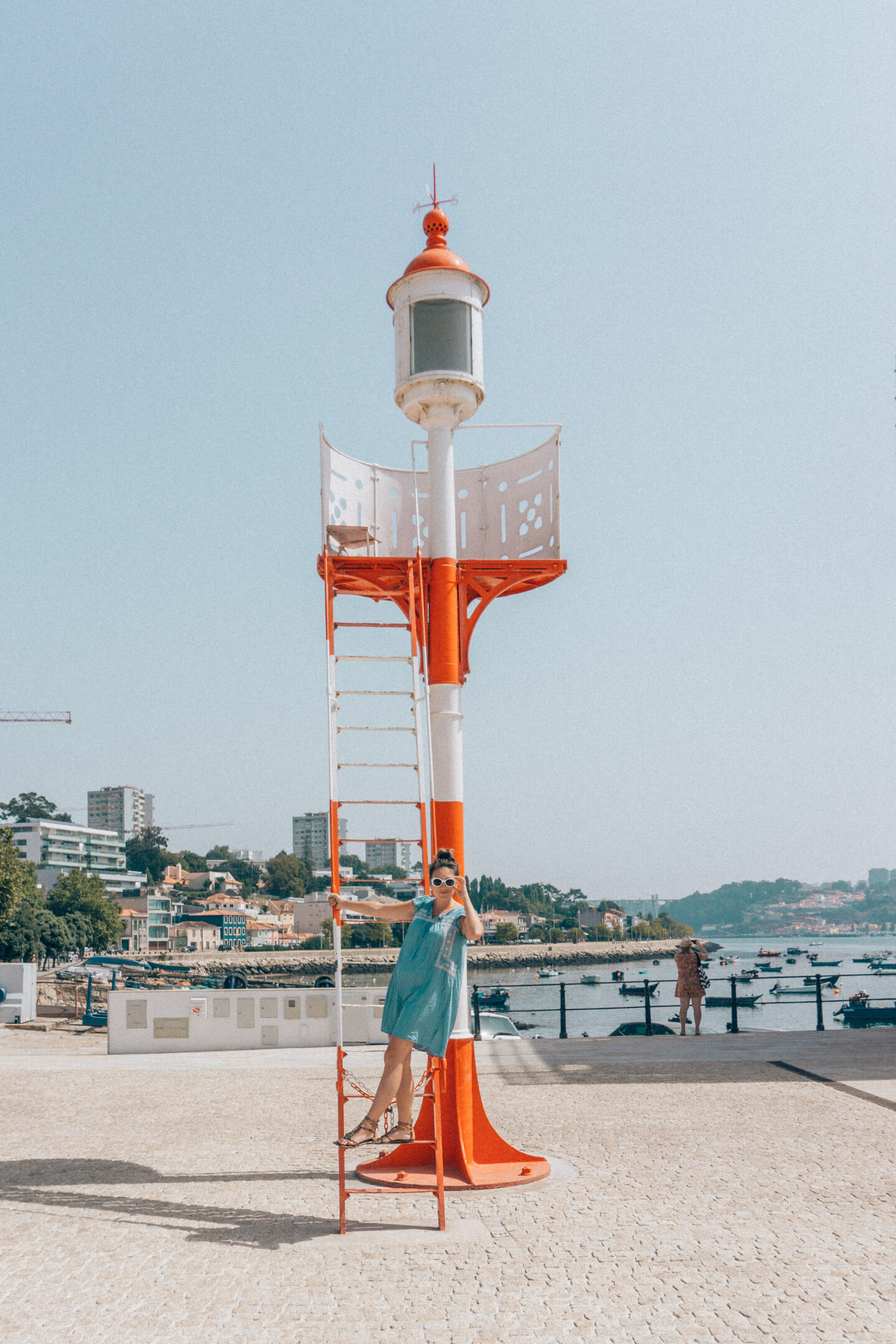 A girl with white sunglasses, hangs off a simple white and red lighthouse in the Foz do Douro beach neighborhood of Porto, Portugal