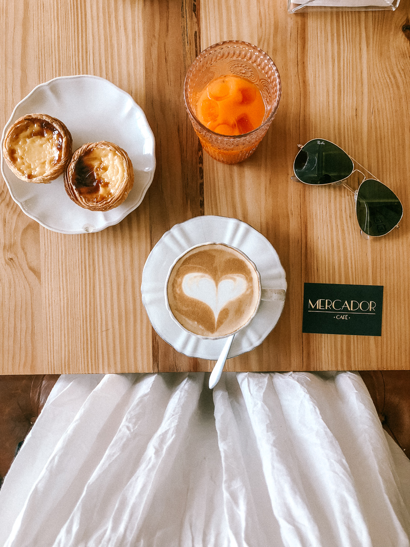 Looking down at a table in Mercador Cafe in Porto. On the table is a  cup of orange juice, sunglasses, a cafe latte, and two Pastel de Natas