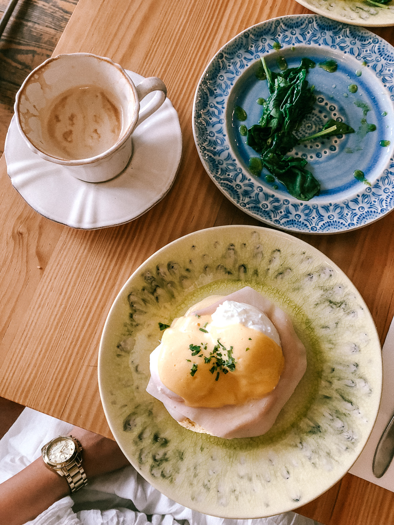 Looking down at a table in Mercador Cafe in Porto. On the table is a cafe latte, and eggs benedict, and a plate of spinach.