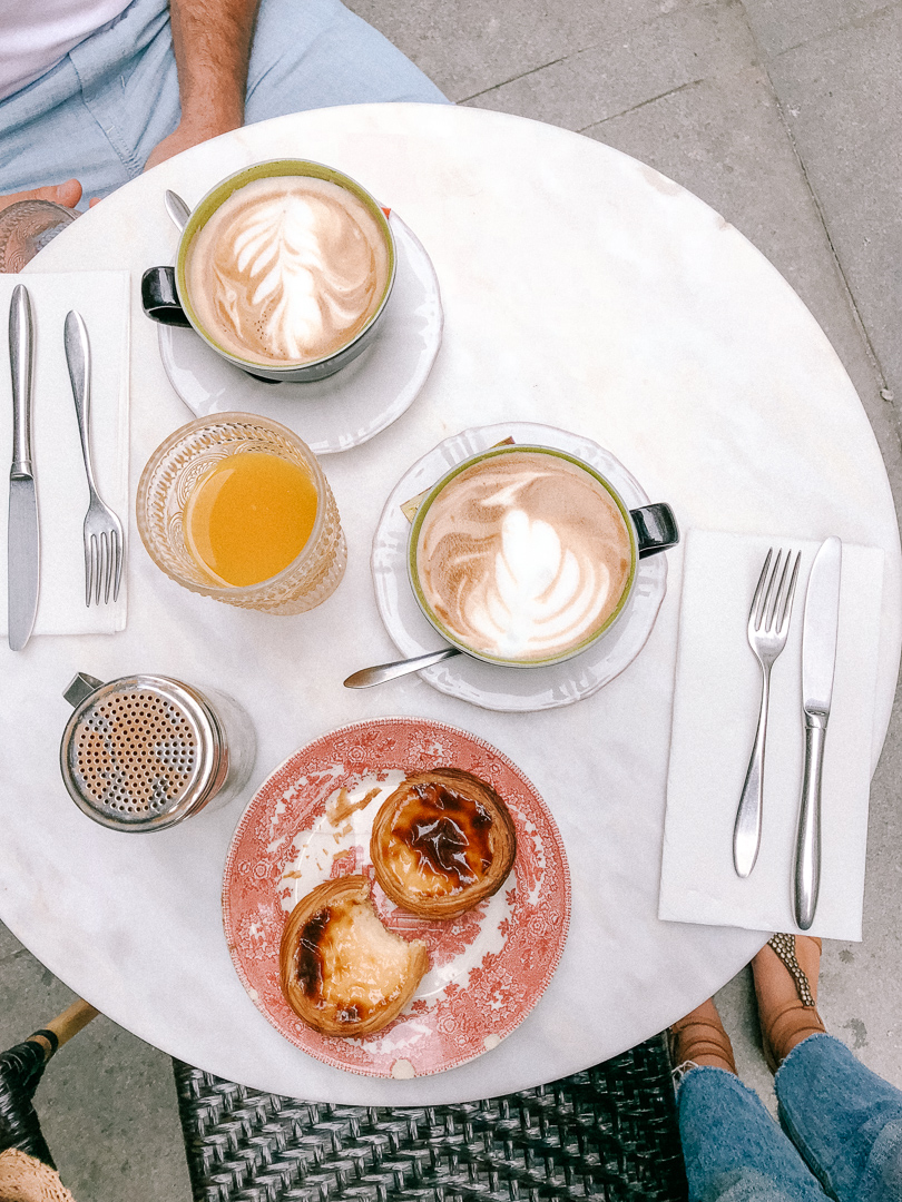 A couple sits at a cafe table in Porto and has two cafe lattes, two Pastel de Natas, and a cup of orange juice