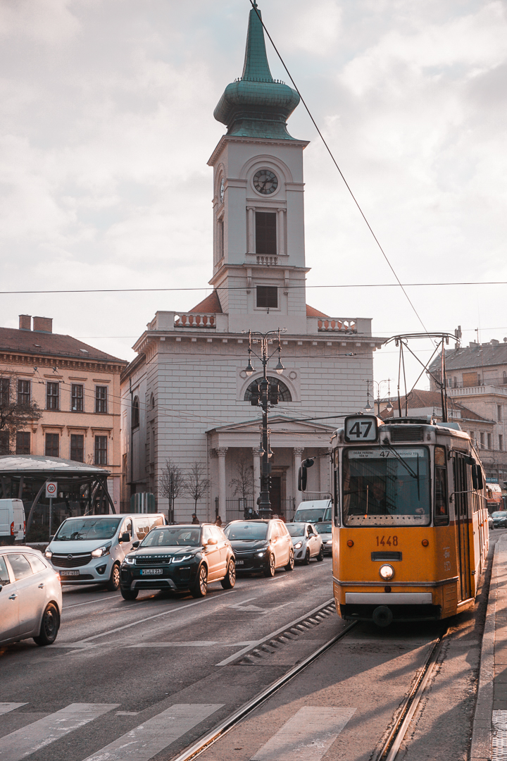 A iconic yellow tram with the number 47 pulls through the streets of Budapest with a church building in the background. 