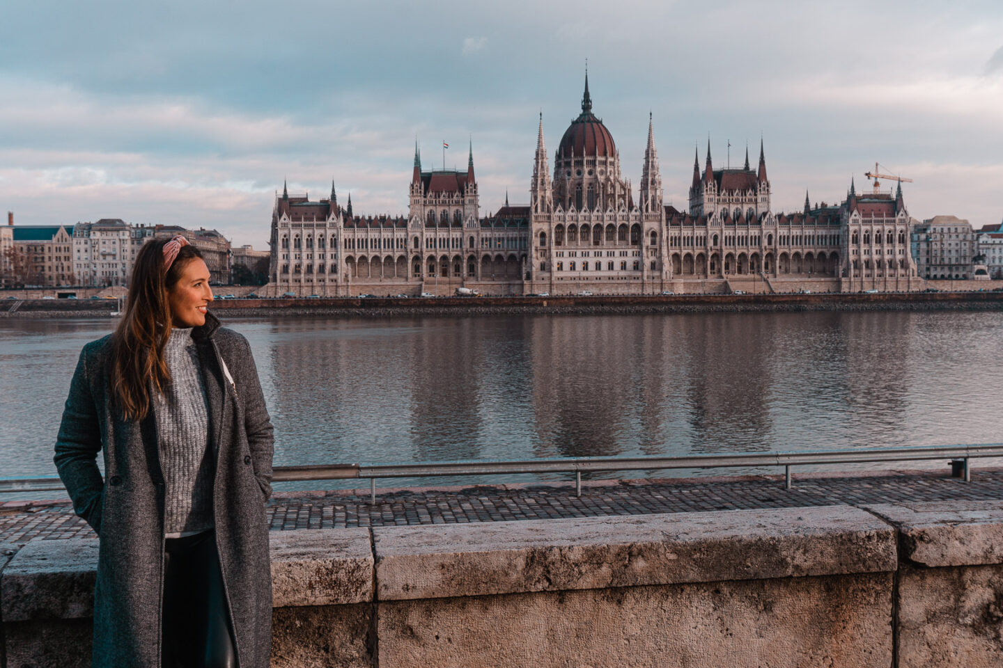 Girl in a winter outfit stands beside the Danube River with the Budapest Parliament house in the background. - 3 Days in Budapest Itinerary