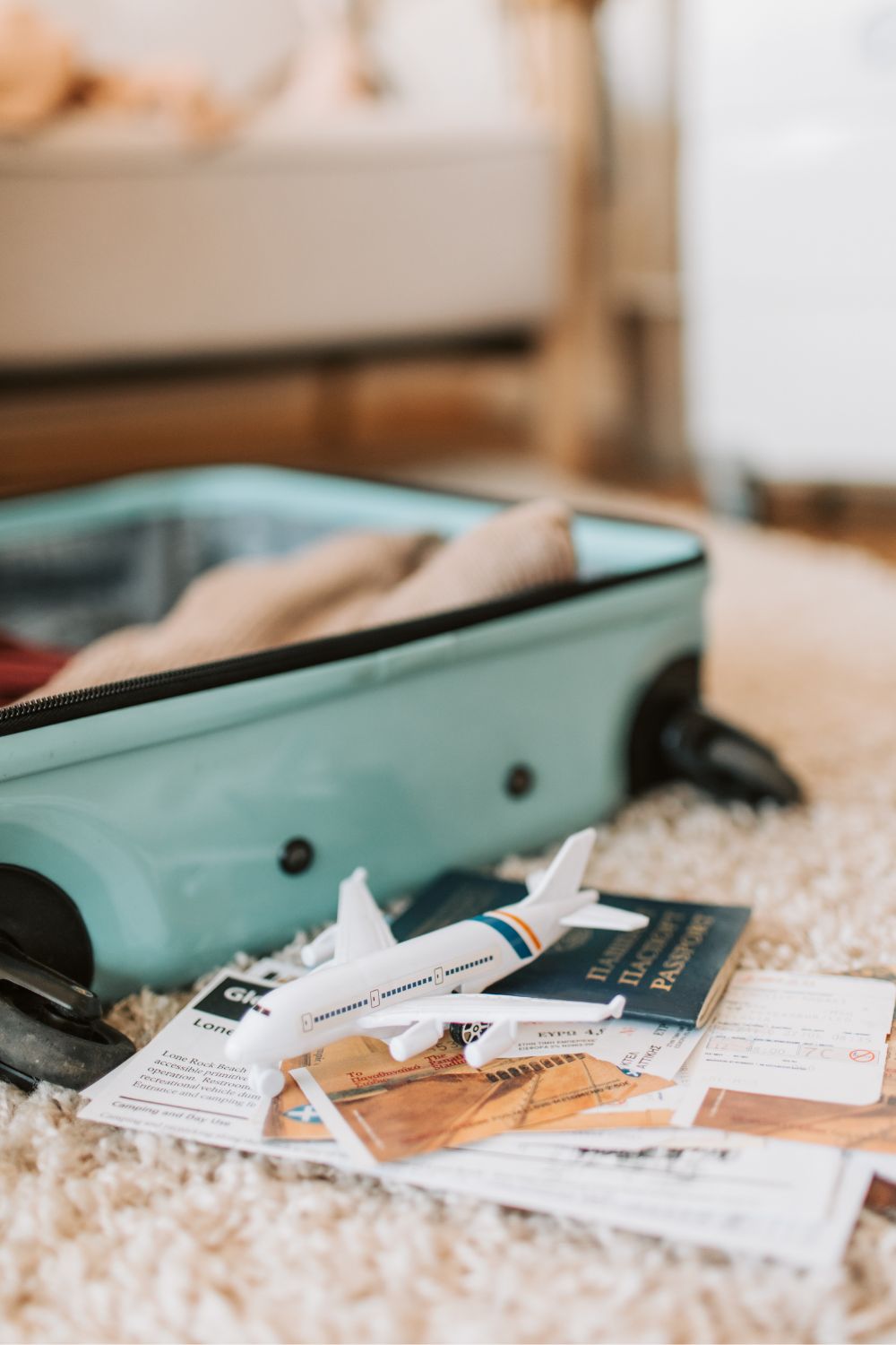 A mint green carry on suitcase lies on a shaggy carpet with a stack of travel documents on the floor beside it and a toy plane on top. 
