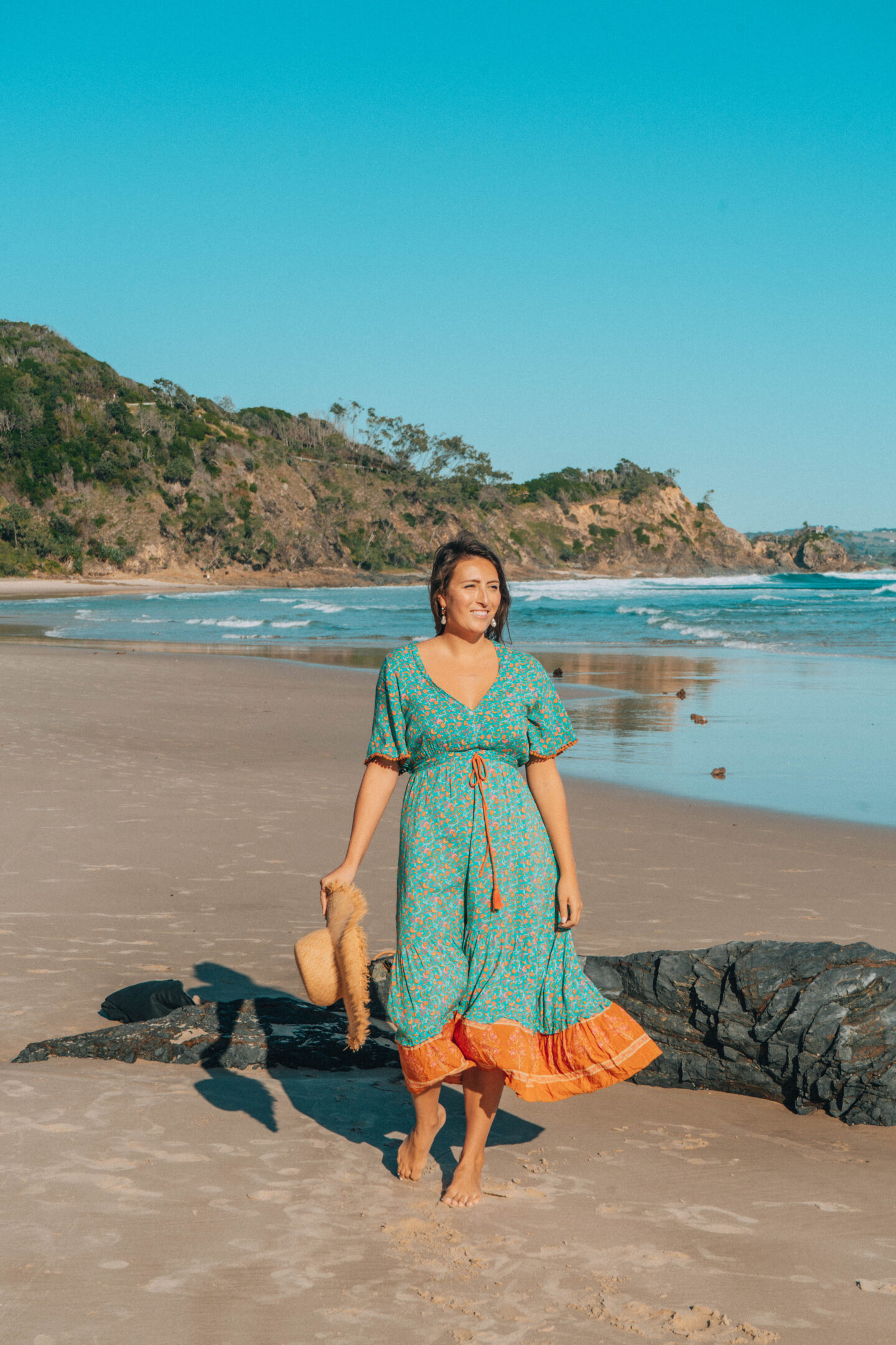 A girl in a green dress holds onto her hat, as she walks along Wategos beach in Byron Bay, Australia.