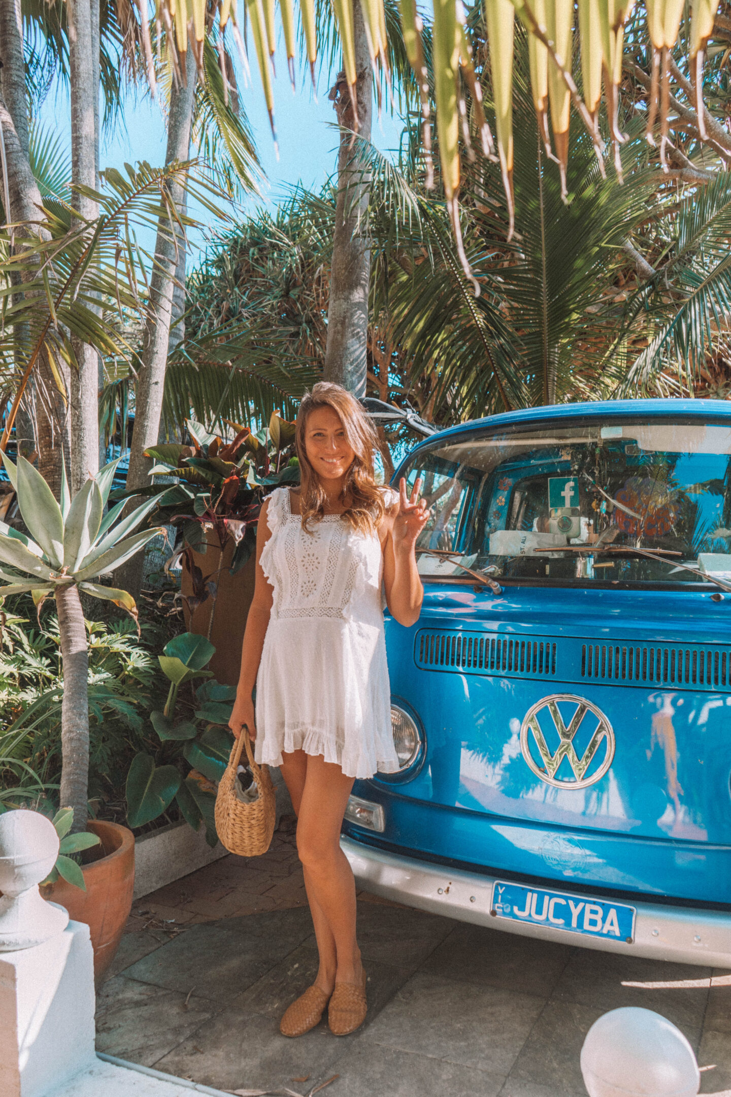 A girl in a white dress puts up the peace sign in front of a blue Volkswagon van in Byron Bay, Australia