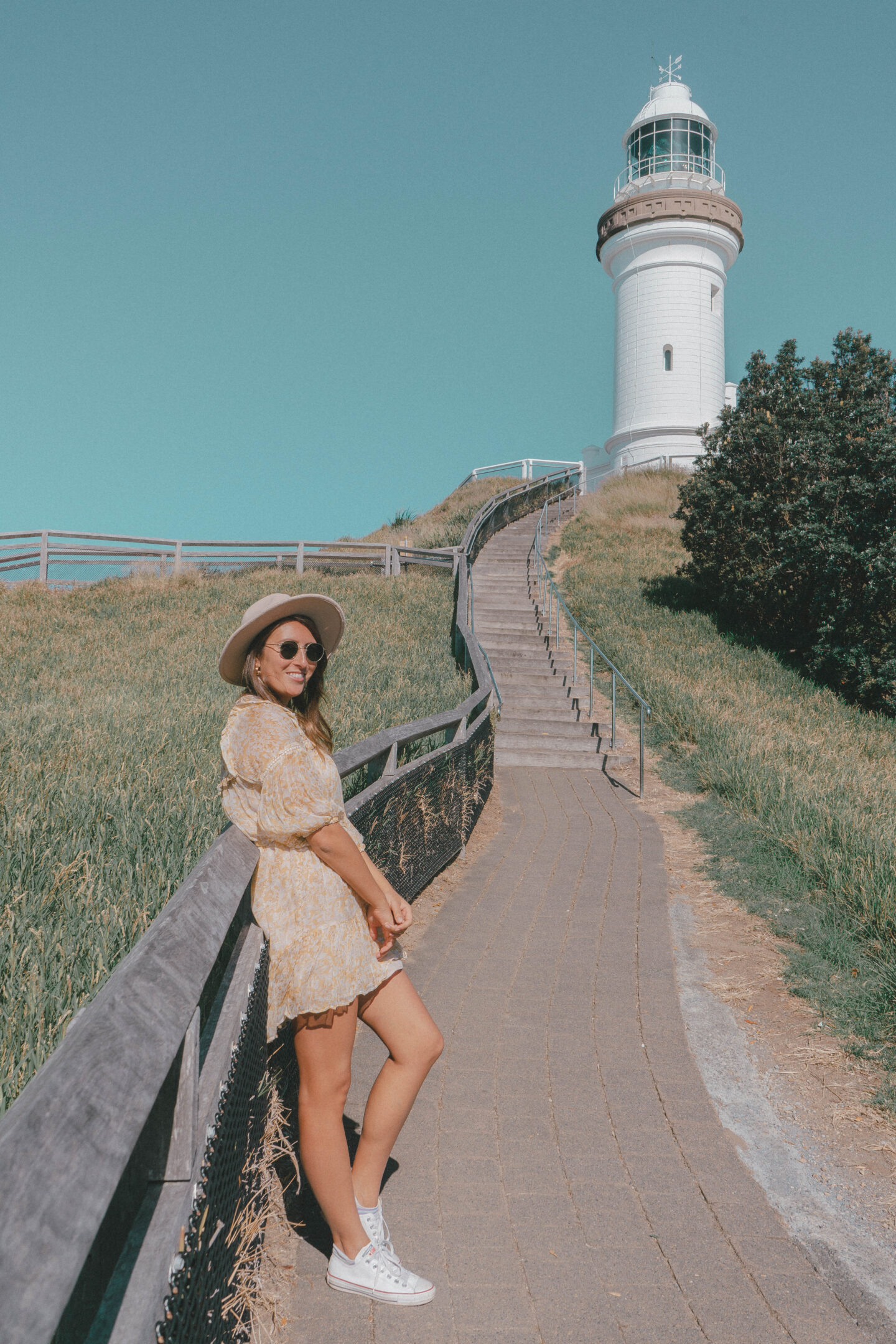 A girl in a yellow dress leans against a fence in front of the iconic Byron Bay white lighthouse that marks the most eastern point in Australia
