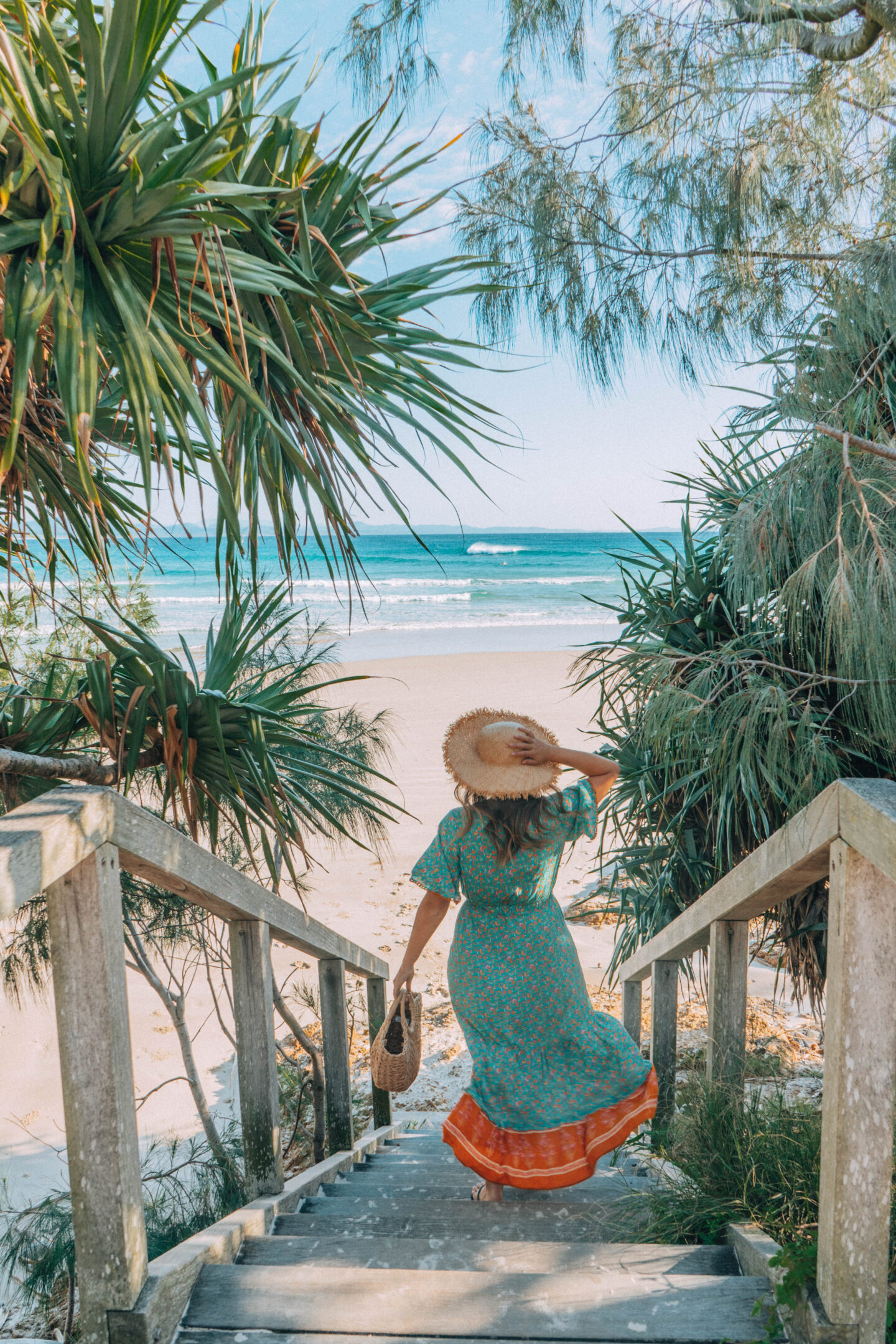A girl in a green dress holds onto her hat as she walks down the stairs to Wategos beach in Byron Bay, Australia.
