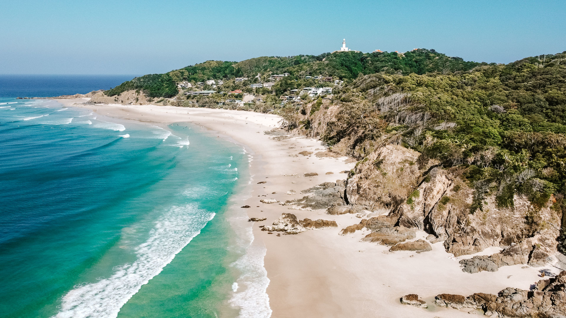 A drone photo looking down to the beautiful blue waves splashing up against Wategos beach during low tide, and the Iconic Byron Bay Lighthouse in the distance. 