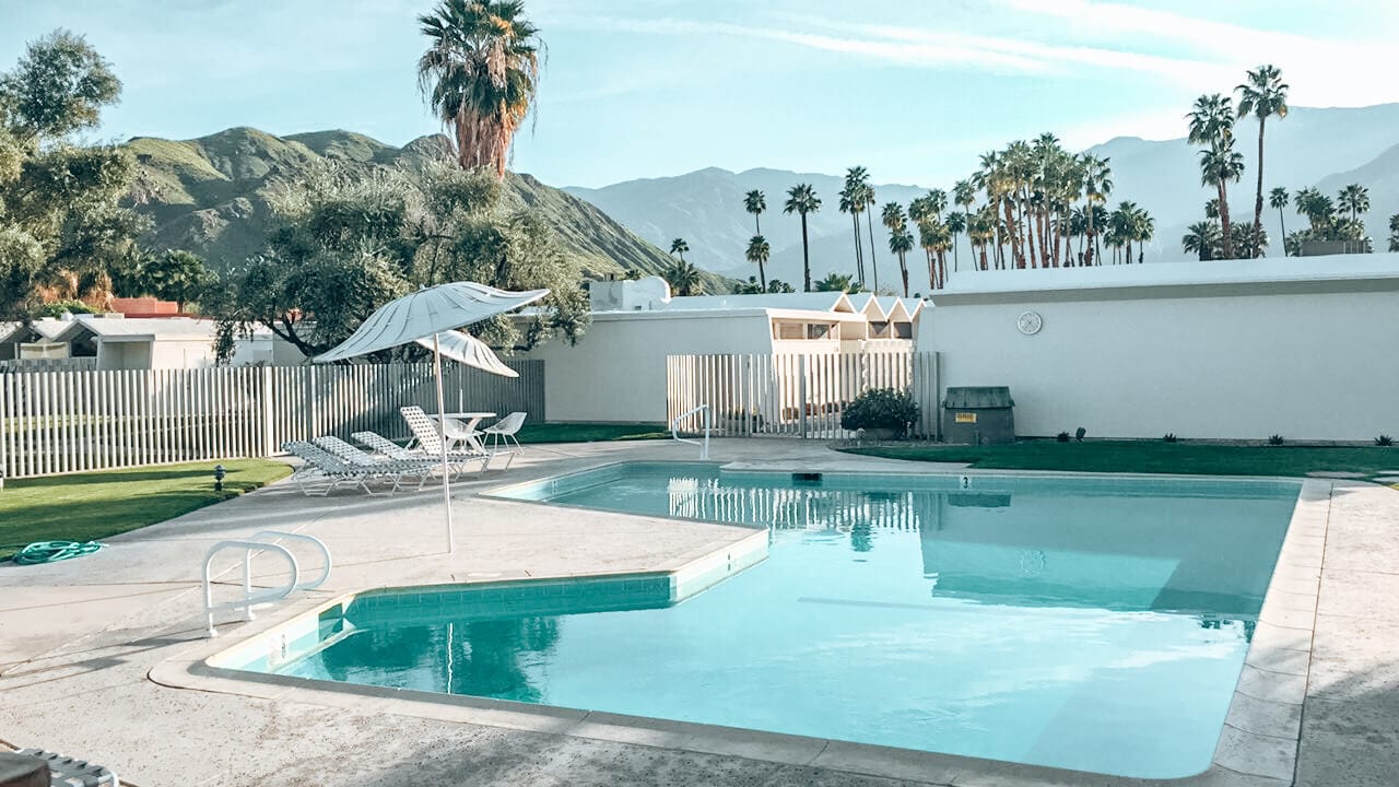 A retro Palm Springs pool surrounded by palm trees, desert mountains and umbrellas
