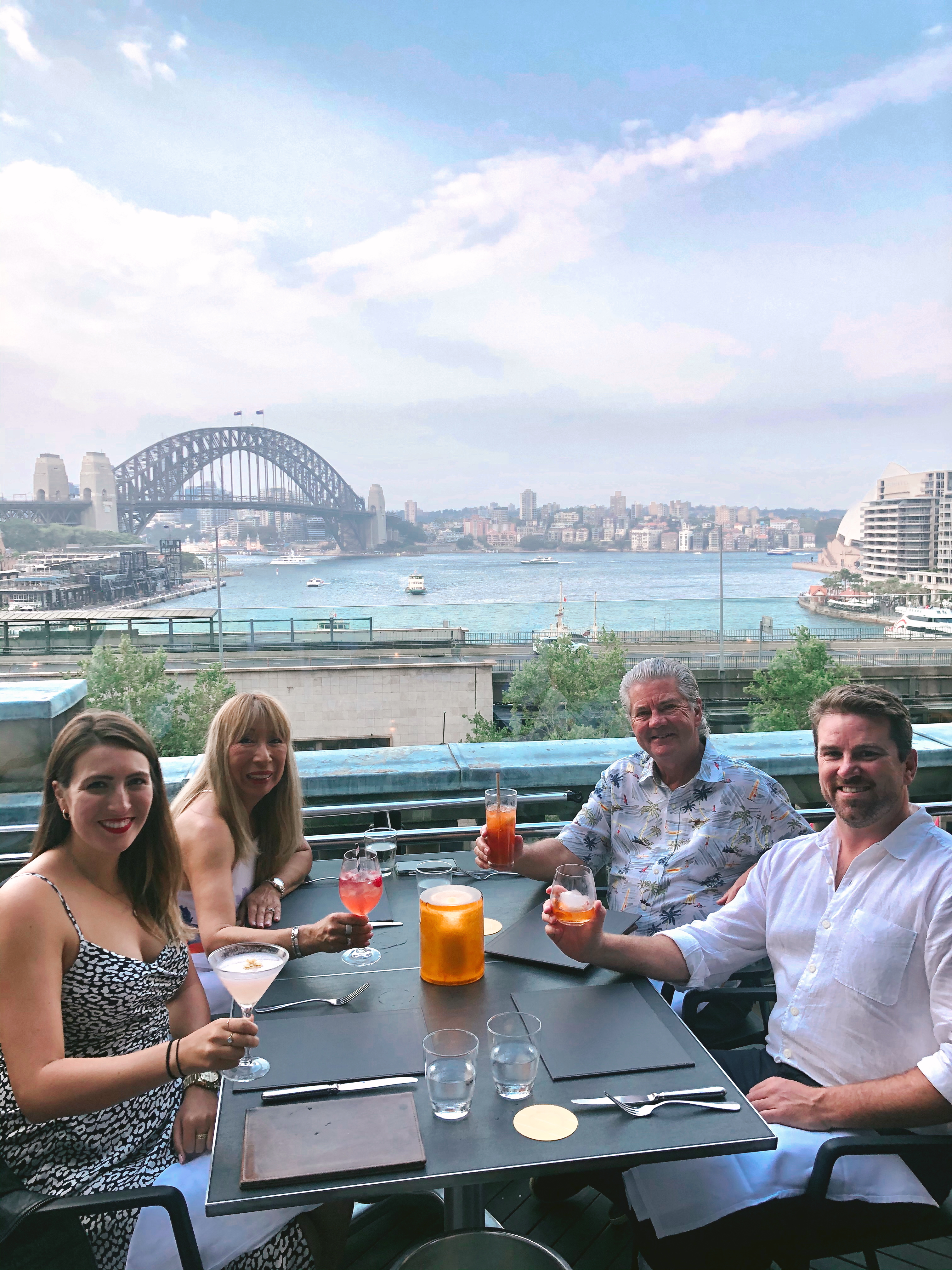 A family of 4 sit at Cafe Sydney with cocktails in hand, and with the iconic Sydney bridge and Opera house in the background.