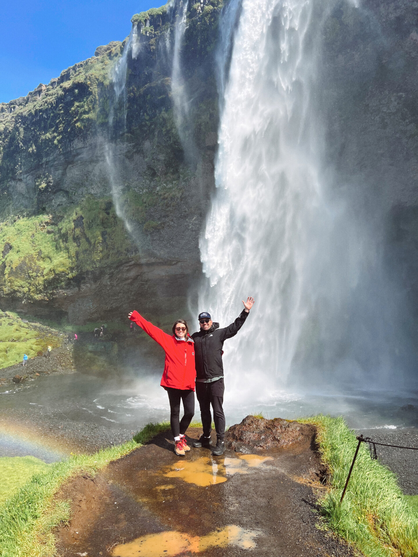 A couple stands in front of the Seljalandsfoss waterfall while mist and rainbows wash over them